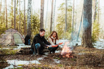 Couple standing outside their tent, warming their hands over a small campfire, with a light dusting of snow on the ground, creating a cozy and warm outdoor camping scene.